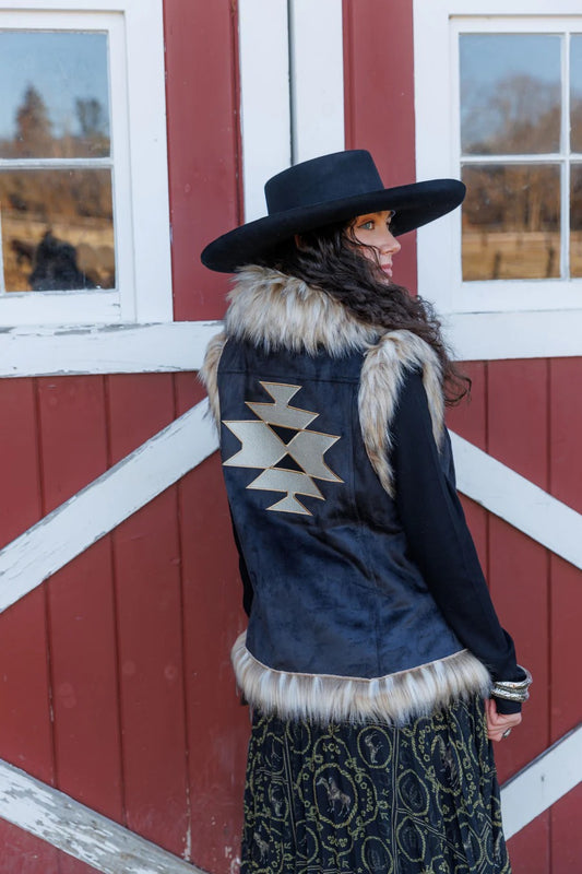 A woman wearing a black jacket with a geometric pattern and fur trim, standing in front of a red barn.