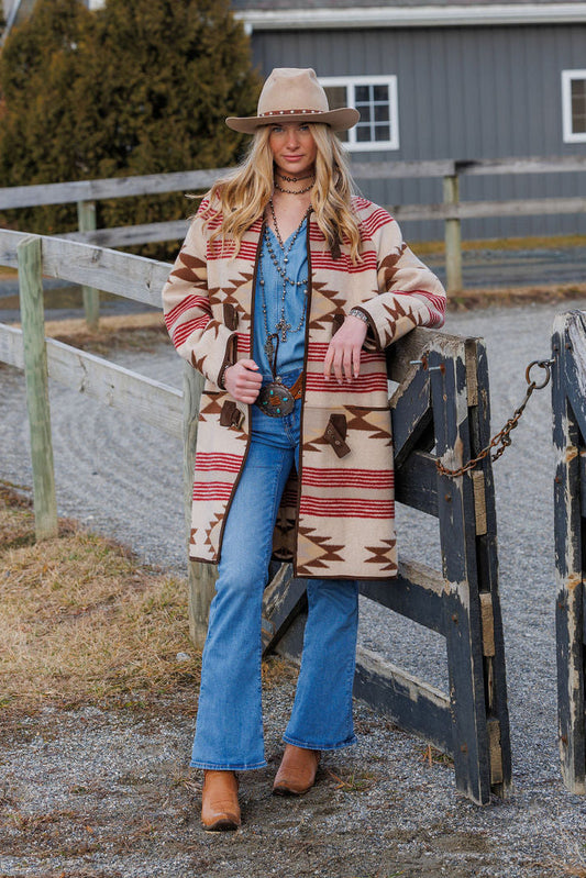 Woman wearing a patterned coat and blue jeans standing on a wooden fence in a rural setting.